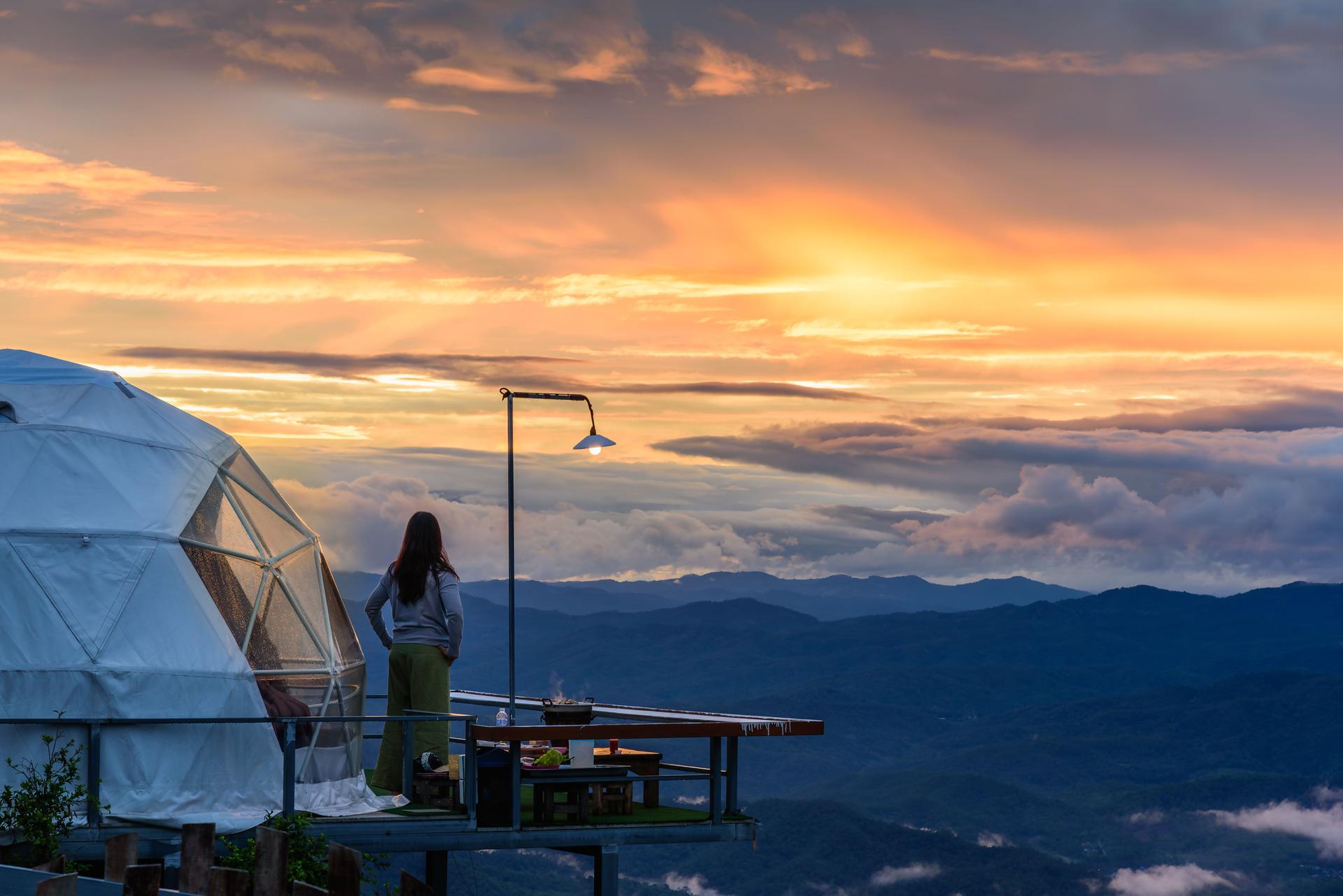 Woman enjoying beautiful sunset sky at glamping dome tent for outdoor camping with mountains view at sunset.